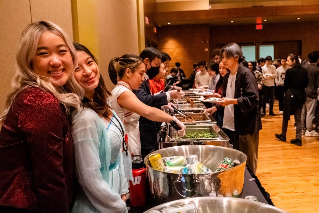 Students smiling in the foreground, with a buffet of food in the background.