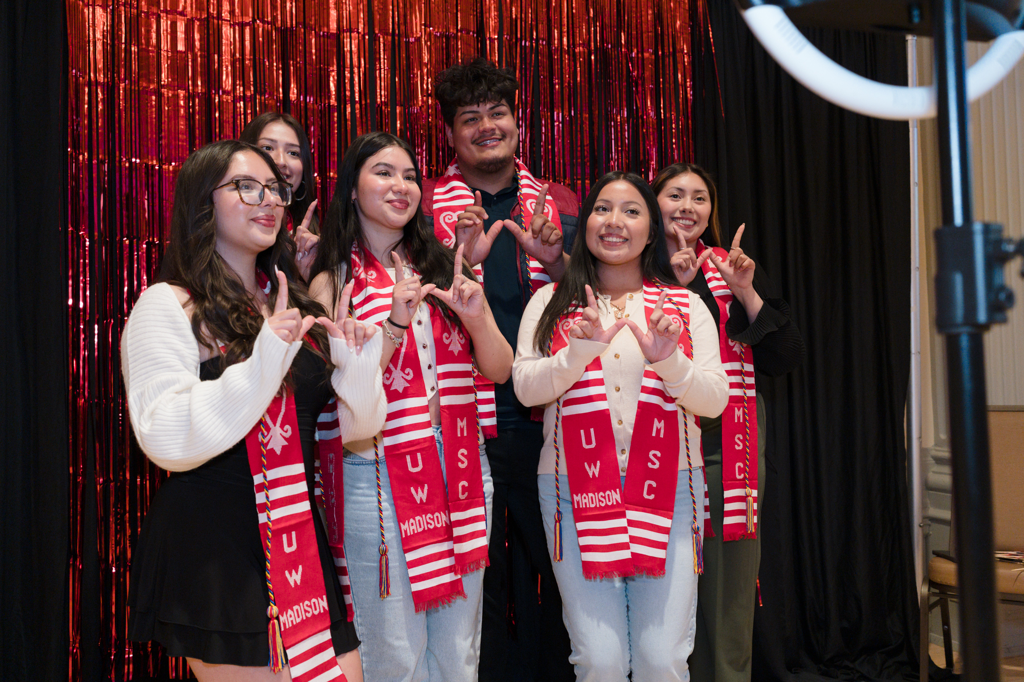 6 students pose with W hands in front of photo booth. all are wearing a red kente cloth stole. 