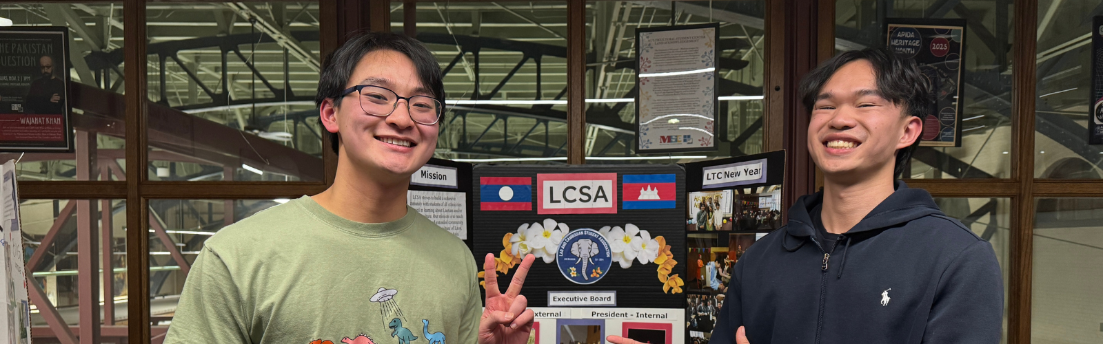 two students smiling at the camera with a student organization poster between them.
